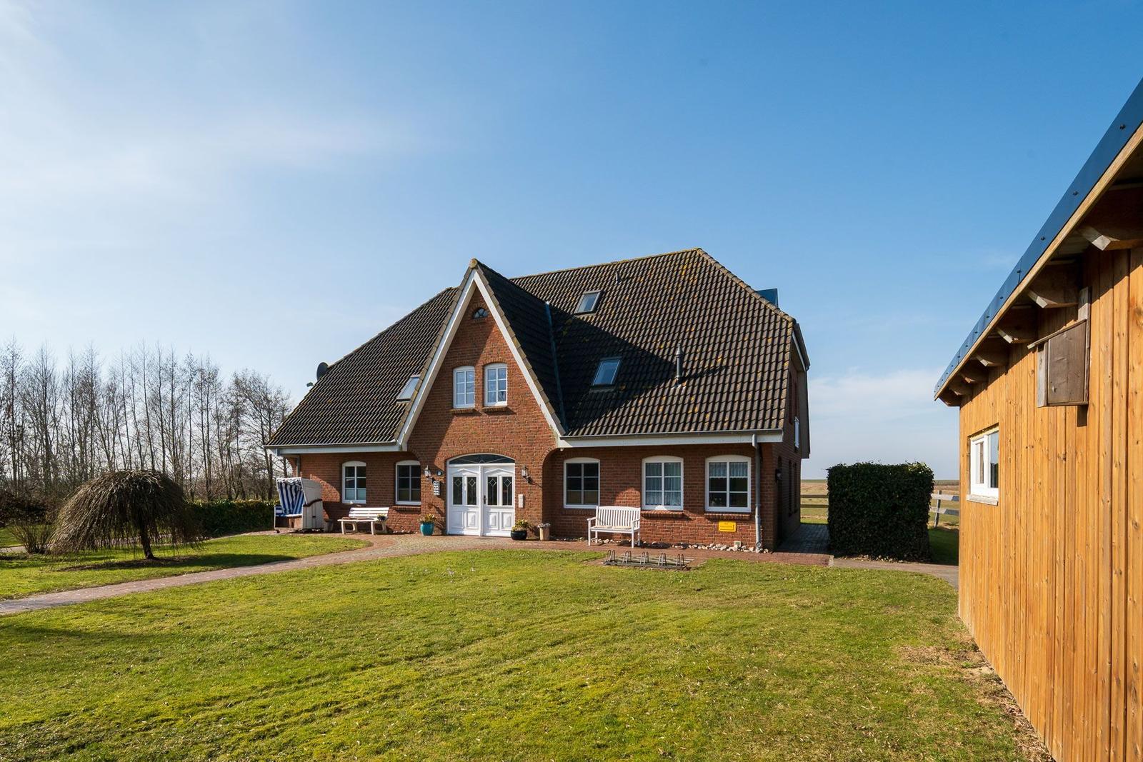 Red brick house with dormers and white front door. Lawn and wooden building to the right.