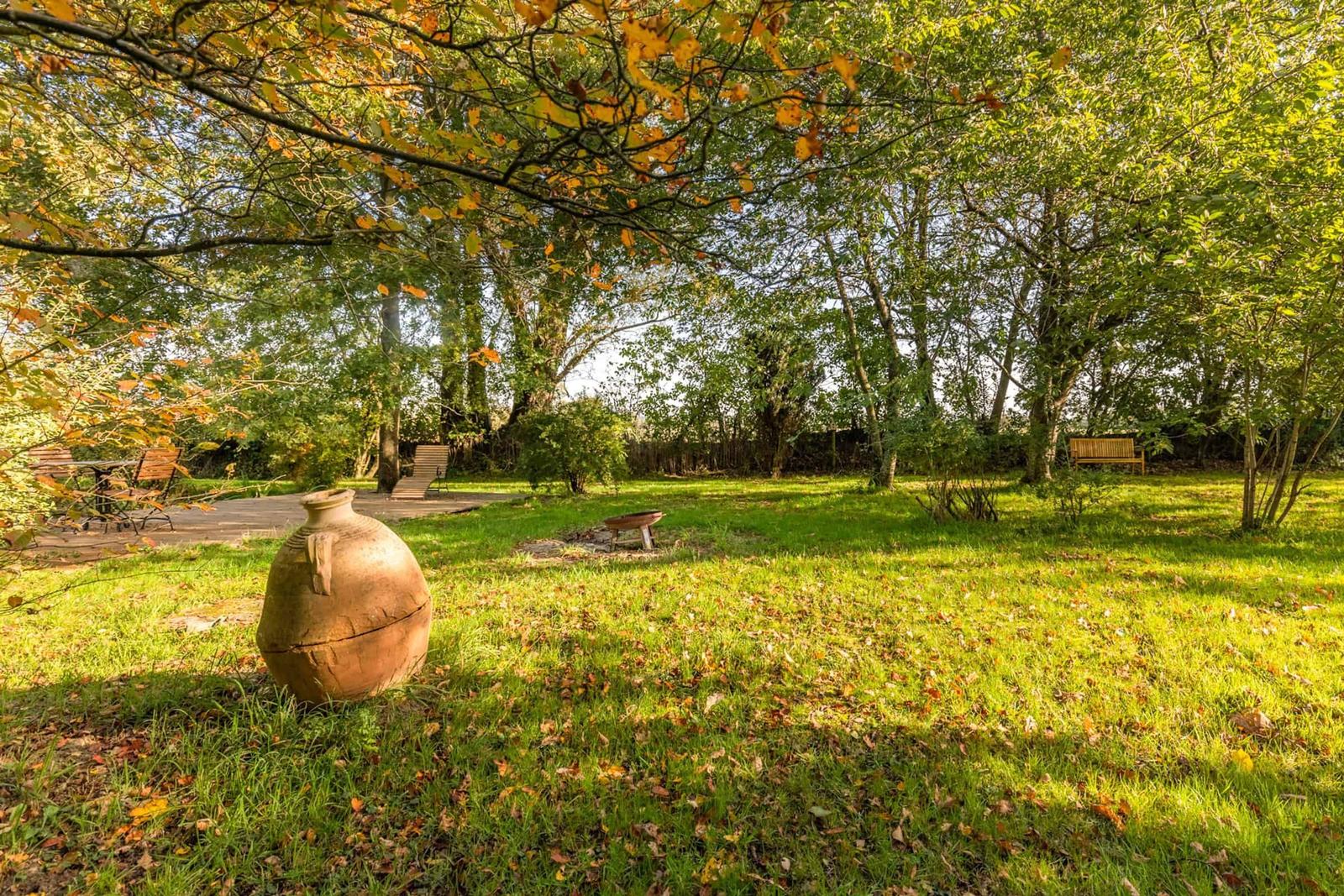 Grüner Garten mit Bäumen, Holzbank und großer Terrakottavase im Vordergrund.