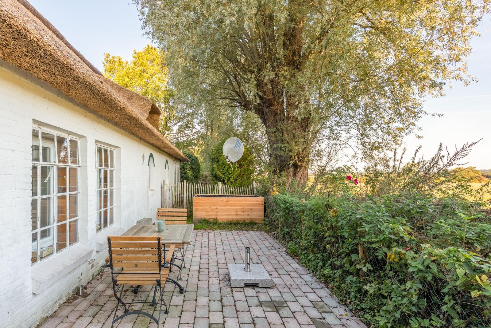 Haus mit Strohdach, Terrasse mit Tisch und Stühlen, große Bäume und Garten.