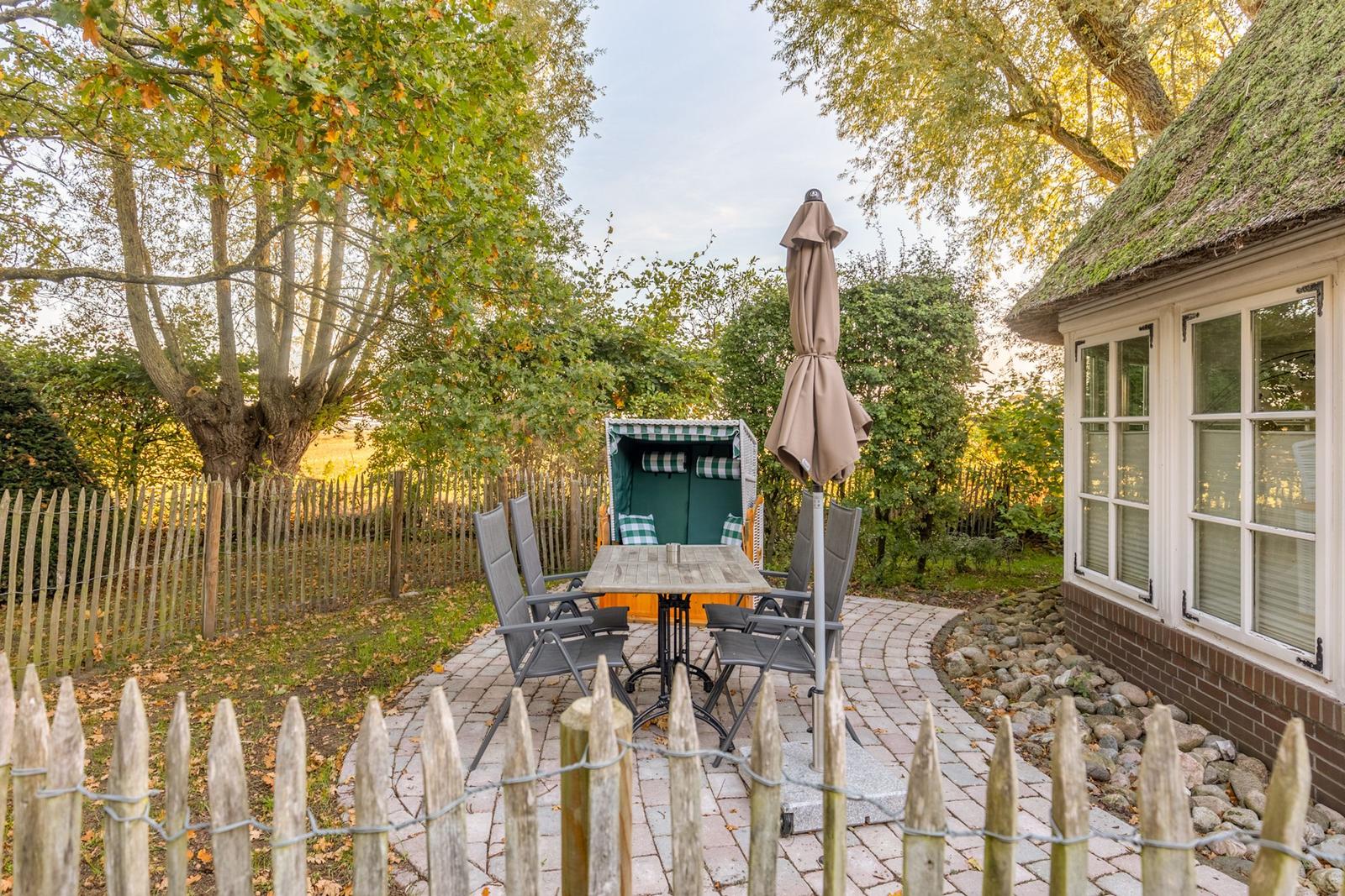 Hinterhof mit Tisch, Stühlen, Sonnenschirm und Strandkorb neben Haus mit Schilfrohrdach.