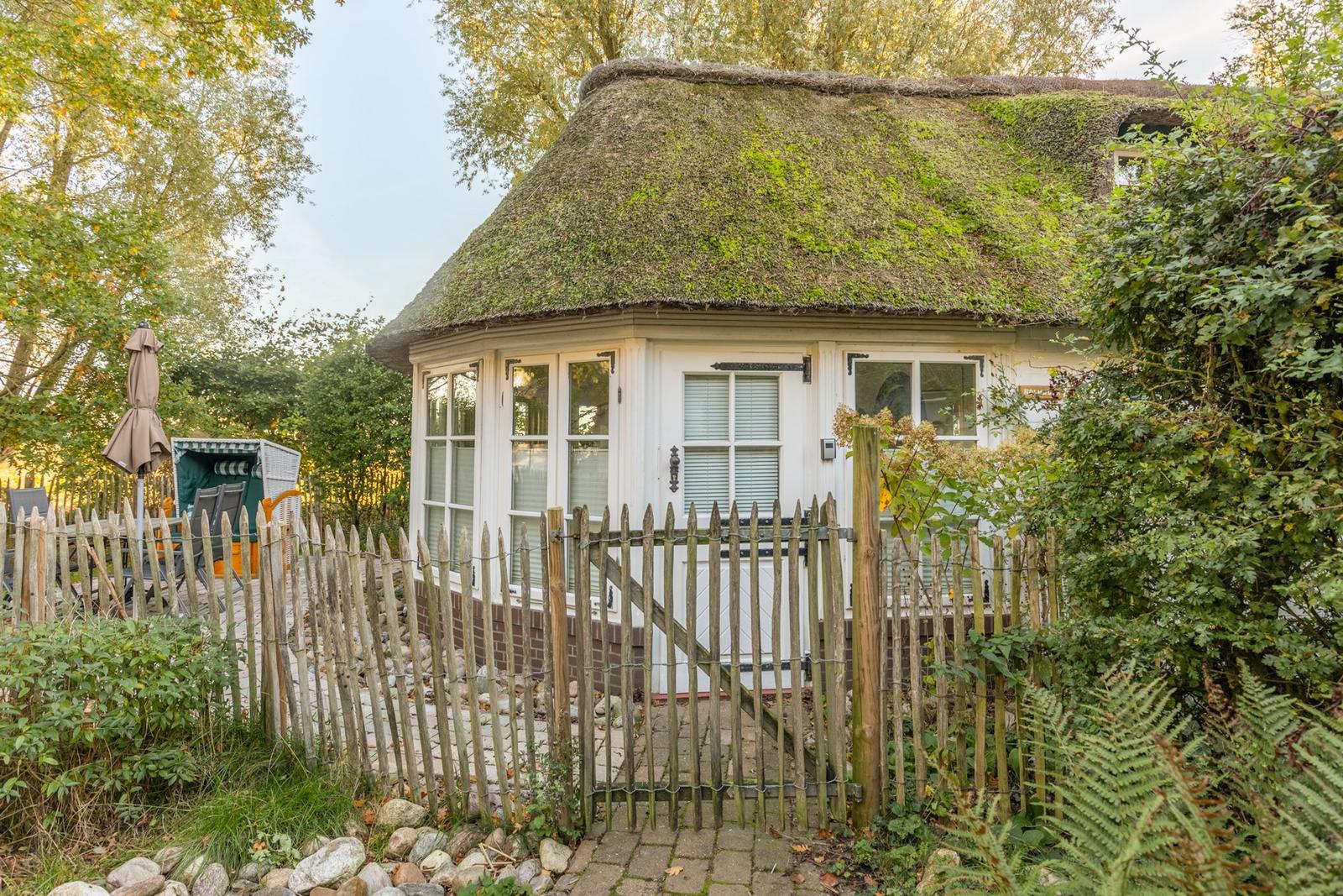 Ein kleines Haus mit Strohdach, umgeben von Grün und einem Holzzaun.