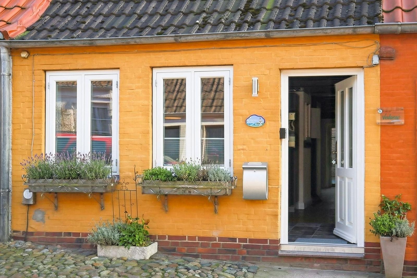 Ein gelbes Haus mit weißen Fenstern und einer offenen Tür. Blumenkästen und ein Briefkasten sind sichtbar.