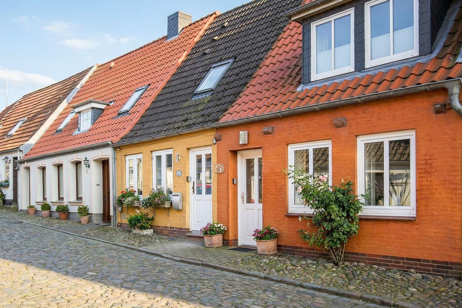 Reihenhaus mit rotem Ziegeldach und weißen Fenstern an einer gepflasterten Straße.