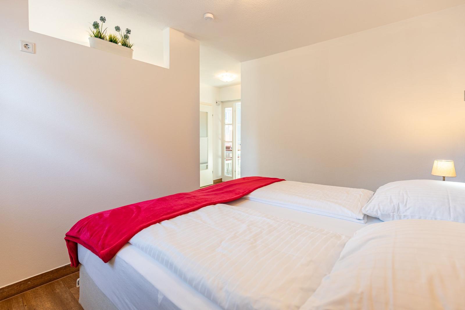 Schlafzimmer mit Bett, roter Decke und Lampe. An der Wand hängt eine Pflanze.
