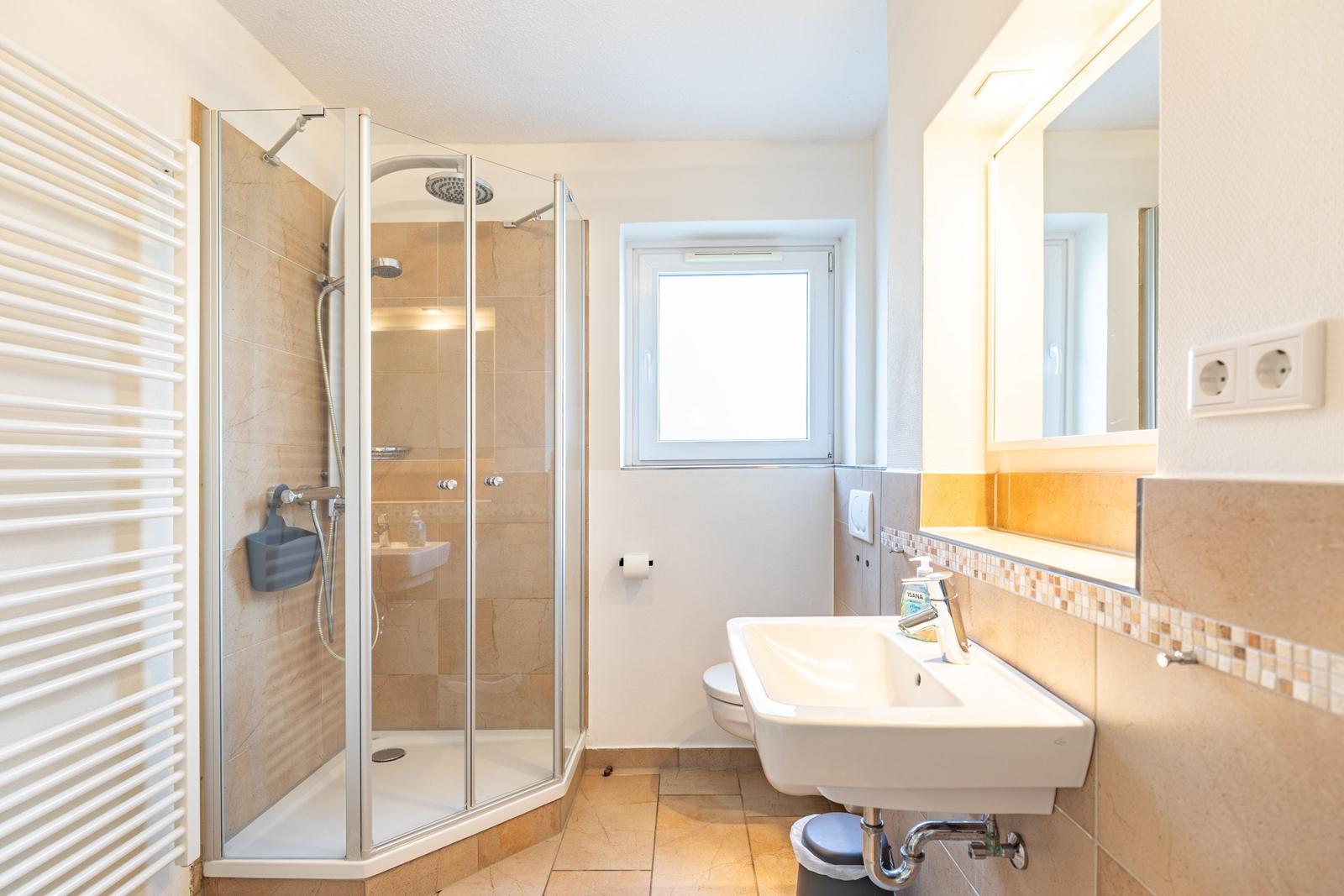 Badezimmer mit Dusche, Waschbecken und Toilette. Fenster sorgt für Tageslicht.