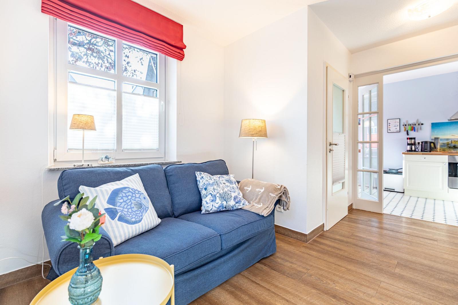 Ein Wohnzimmer mit blauem Sofa, rotem Fensterrollo und Holzboden. Küche ist im Hintergrund sichtbar.