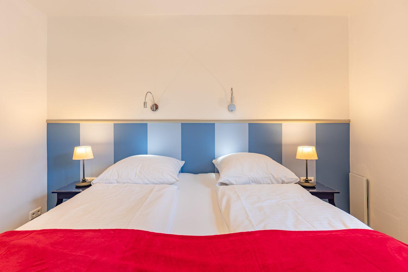 Doppelbett mit rotem Deckenrand und blau-weißem Kopfteil. Zwei Nachttische mit Lampen.