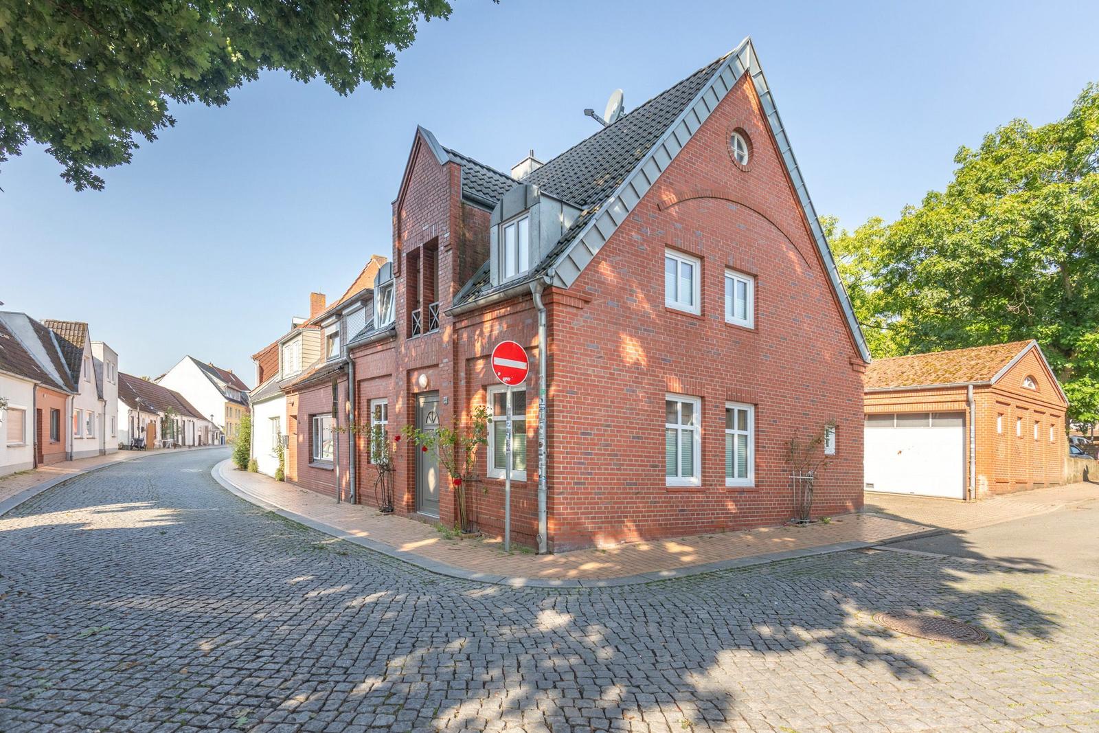 Rotes Backsteinhaus mit Dachgaube und Garagenbereich an einer Straße mit Kopfsteinpflaster.