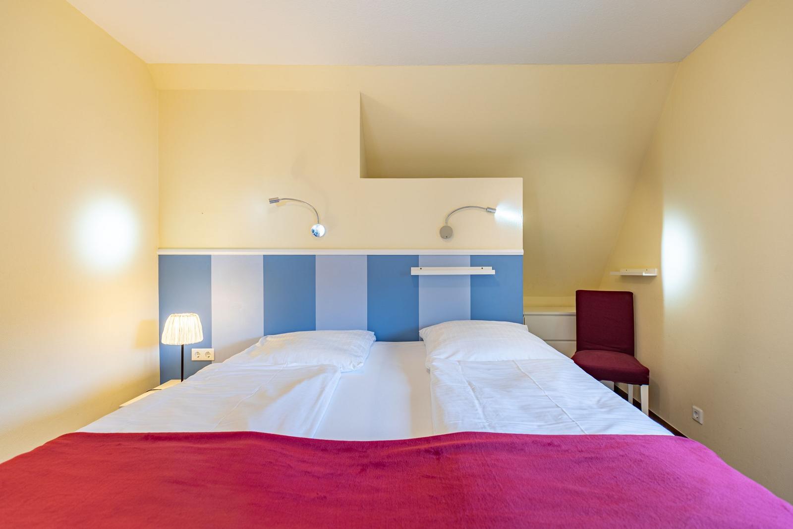 Ein Bett mit weißer Bettwäsche und roter Decke. Blauer Kopfteil und roter Stuhl.