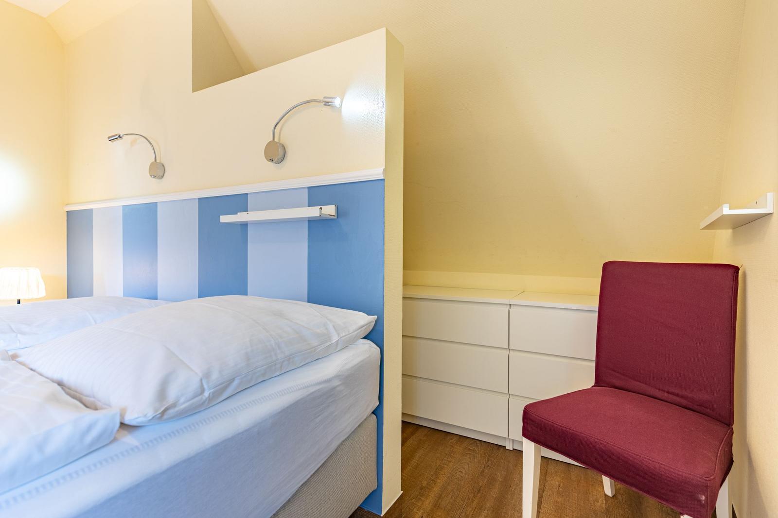 Schlafzimmer mit Bett, blau-weißem Kopfteil, roter Sitzgelegenheit und weißen Schränken.