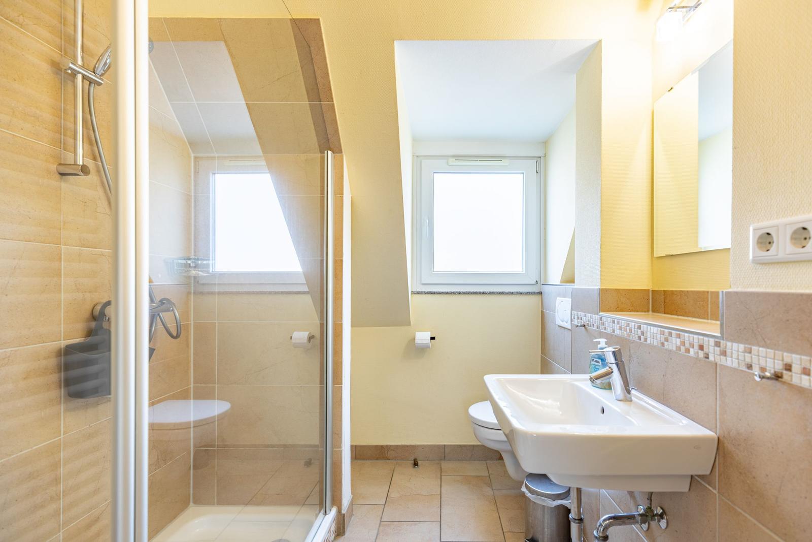 Bad mit Dusche, Waschbecken und Toilette. Fenster sorgen für Tageslicht.