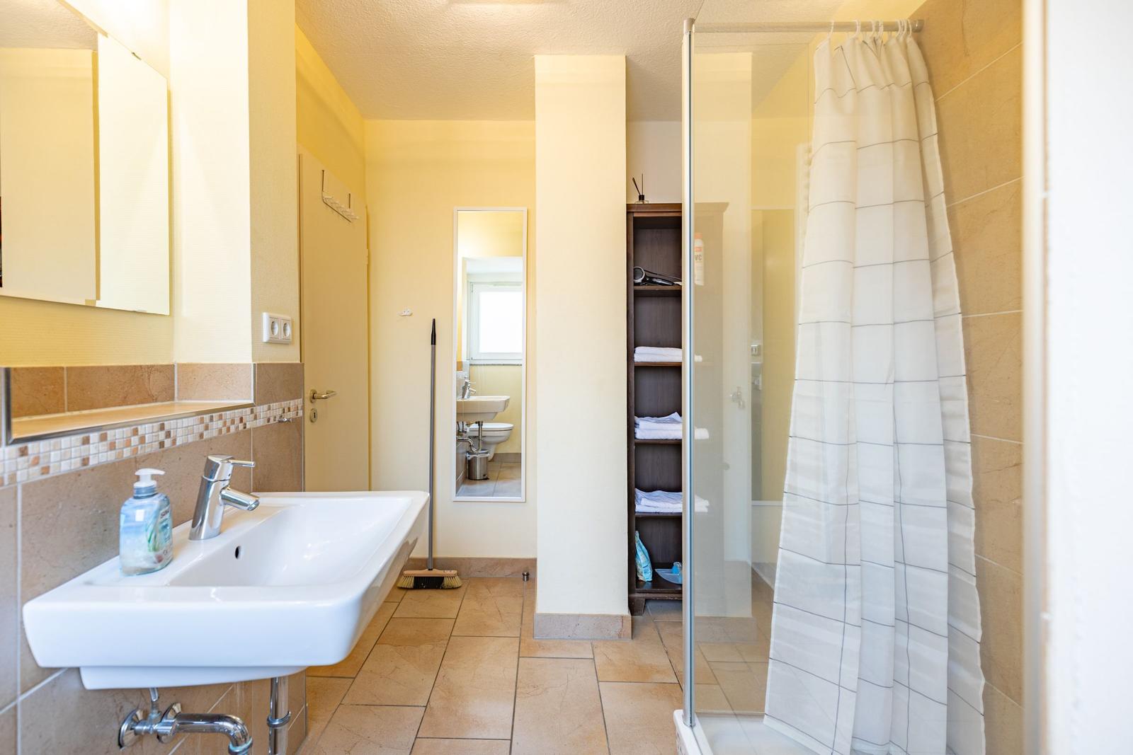 Badezimmer mit Dusche, Waschbecken und Spiegel. Holzregal und Fliesenboden.