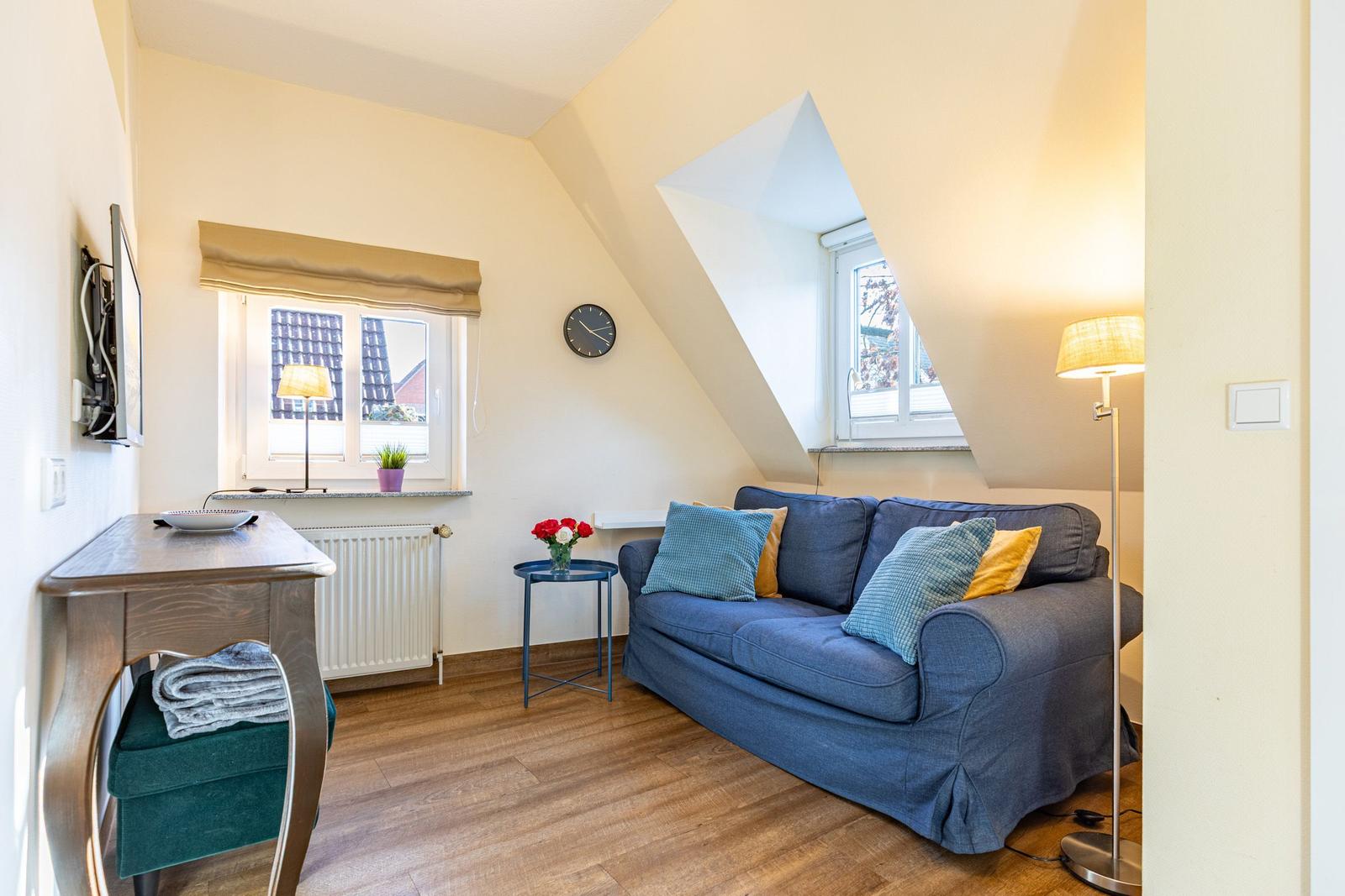 Ein Wohnzimmer mit blauem Sofa, Holzboden und schrägen Wänden. Fenster und Lampen sind sichtbar.