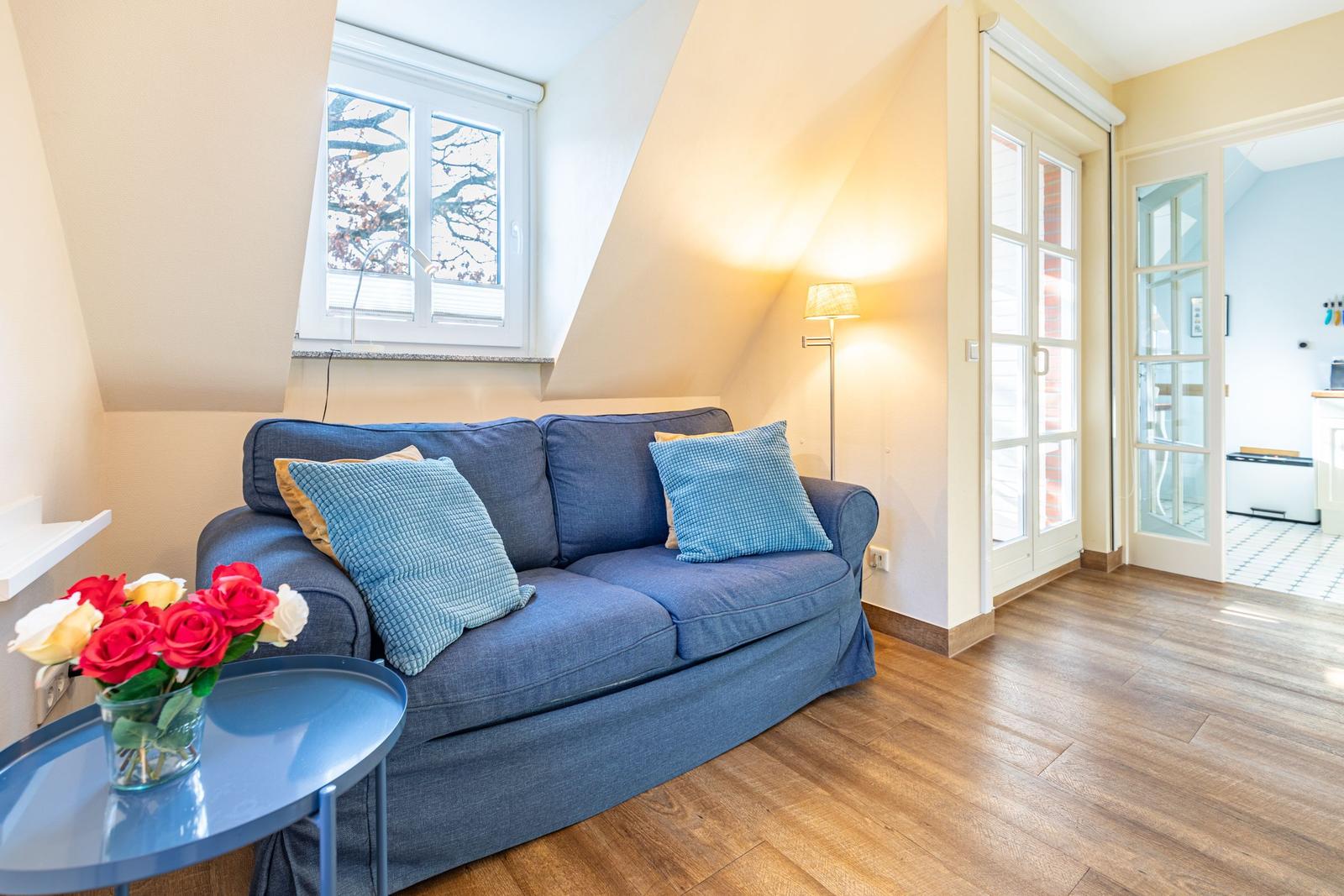 Ein Wohnzimmer mit blauem Sofa, Holzboden und Fenster. Eine Tür führt in die Küche.