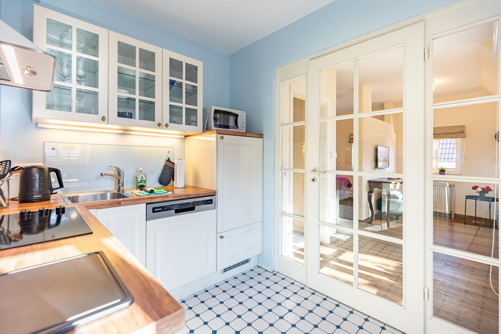 Küche mit weißen Schränken, Holzoberflächen und Fliesenboden. Durchgehende Glastüren führen in das Wohnzimmer.