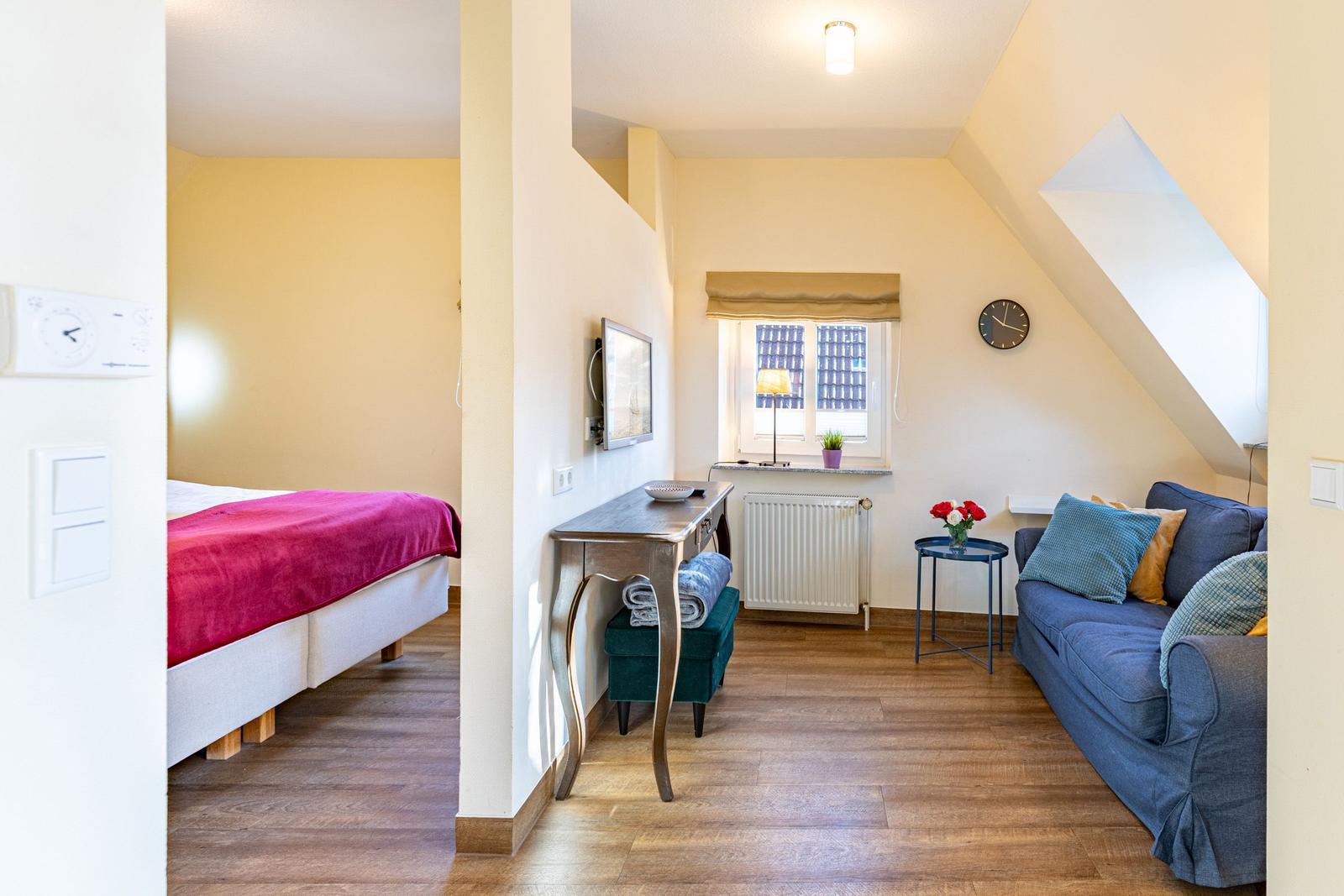 Wohnbereich mit Sofa, Schreibtisch und Bett im Hintergrund. Holzboden und schräge Decke.