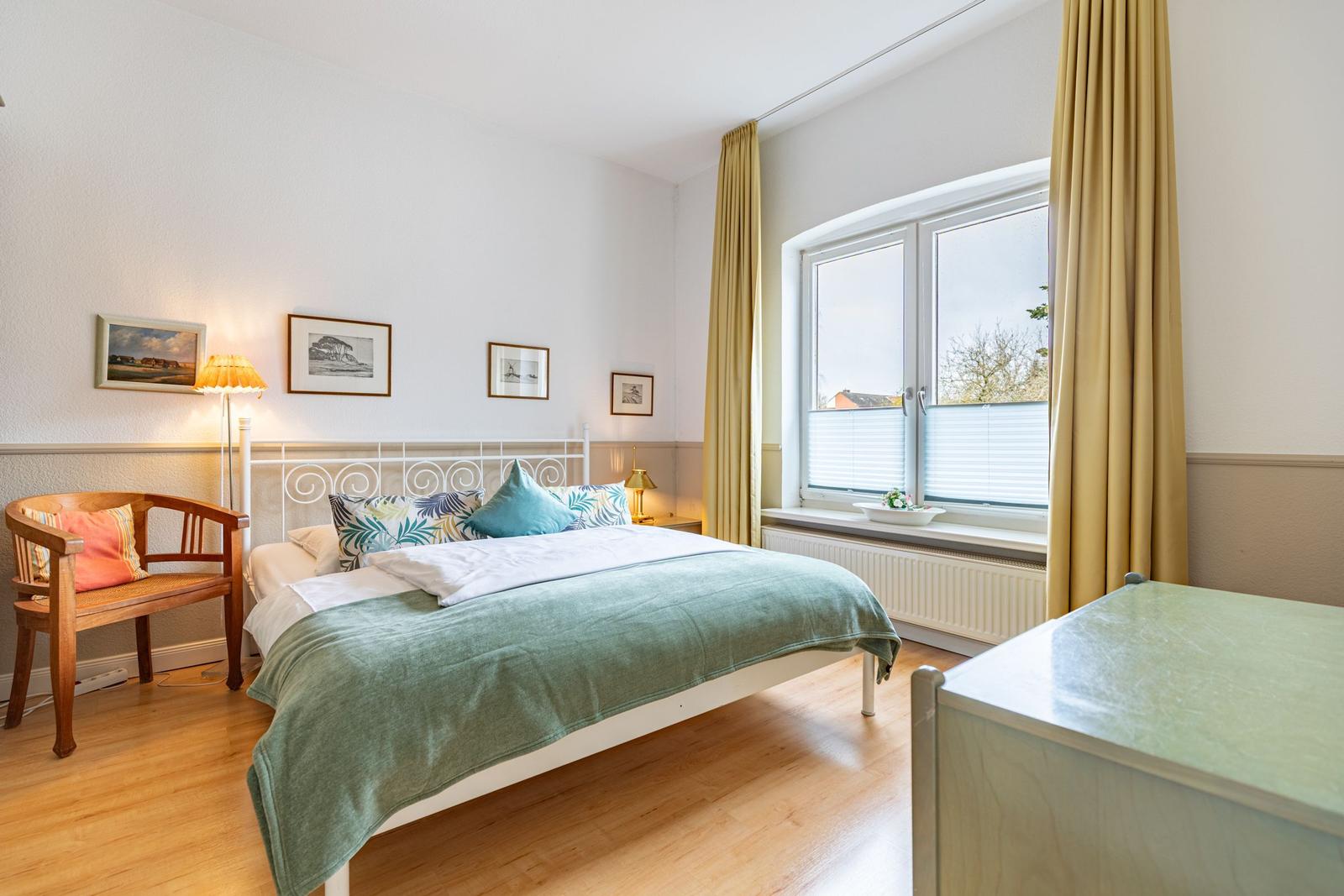 Schlafzimmer mit Bett, Holzstuhl und Fenster mit Vorhängen.