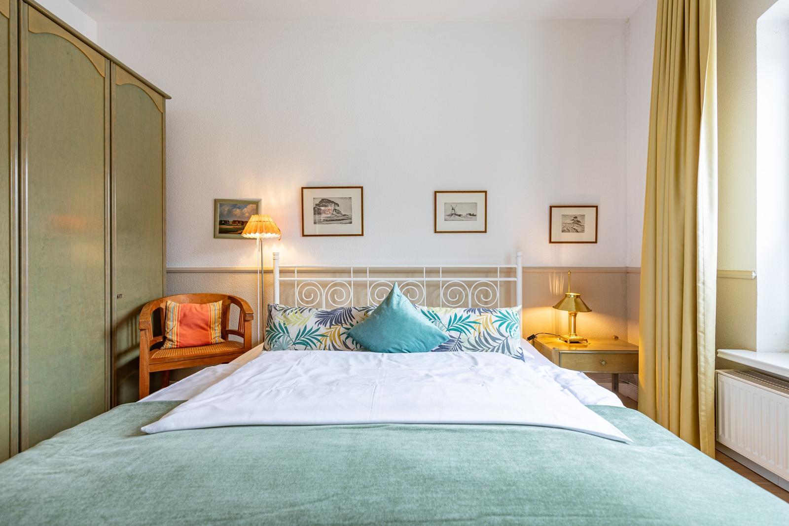 Ein Schlafzimmer mit Bett, Nachttisch, Stuhl und Kleiderschrank. An der Wand hängen Bilder.