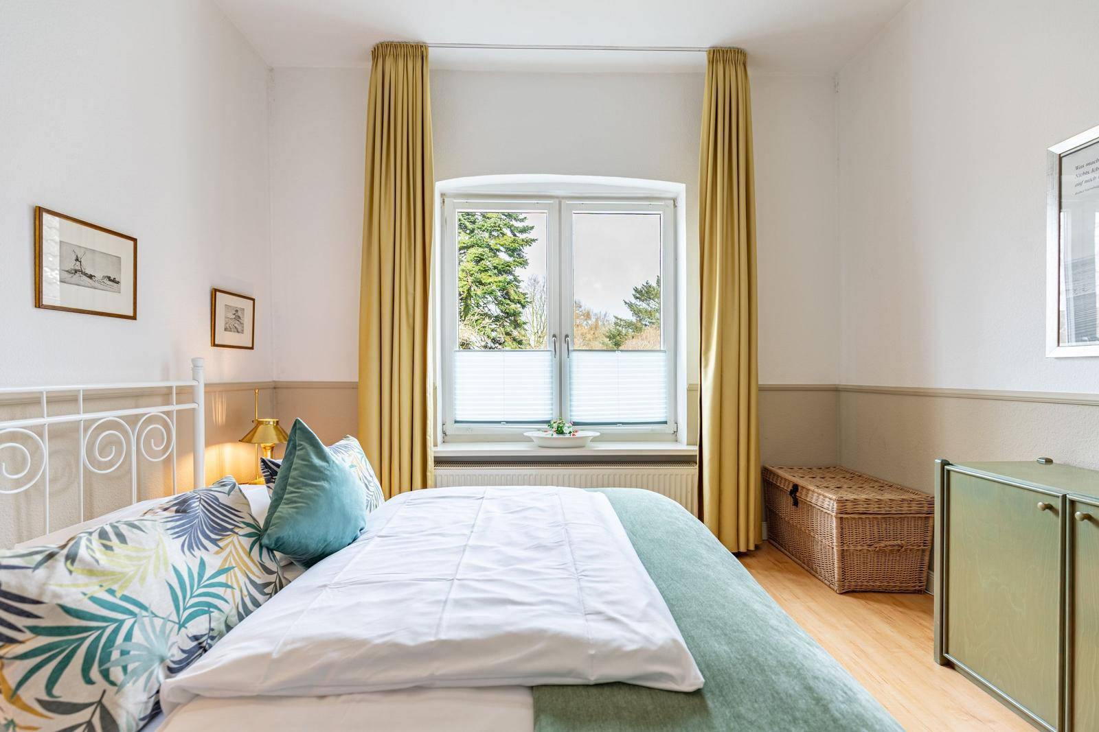 Schlafzimmer mit Bett, Fenster und gelben Vorhängen. Holzboden und Schrank.