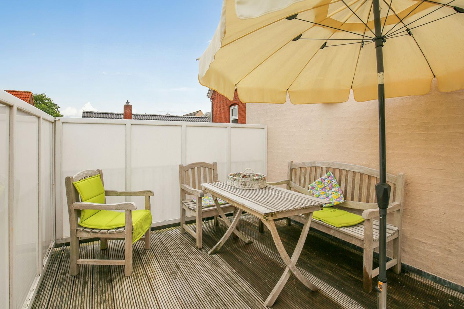Terrasse mit Holztisch, Bänken und gelbem Sonnenschirm. Holzdielenboden und weiße Umzäunung.