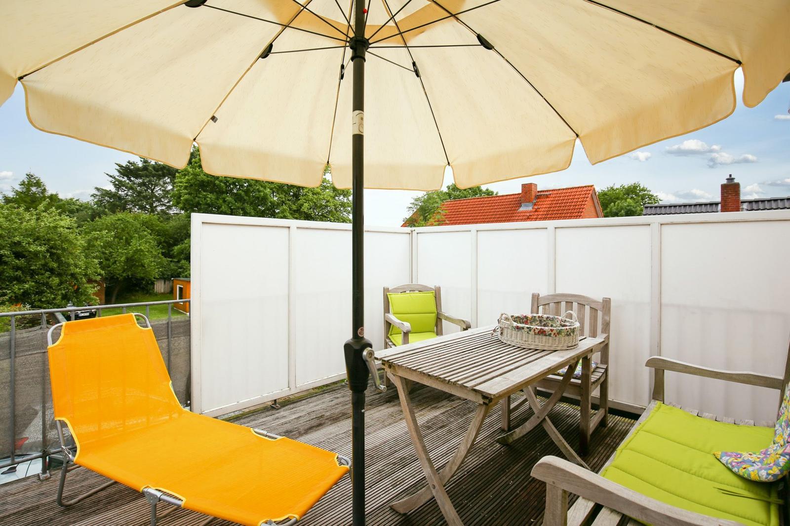 Terrasse mit Sonnenschirm, Holztisch, Stühlen und Liegestuhl. Weiße Umzäunung und Bäume im Hintergrund.