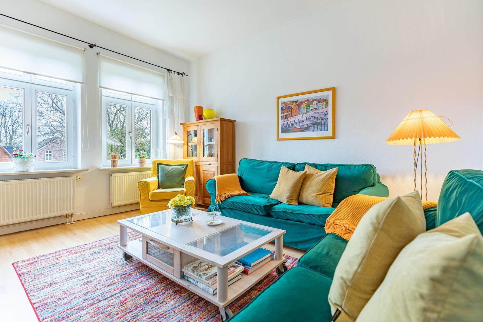 Ein Wohnzimmer mit grünem Sofa, gelber Sessel und Glas-Tisch auf buntem Teppich.