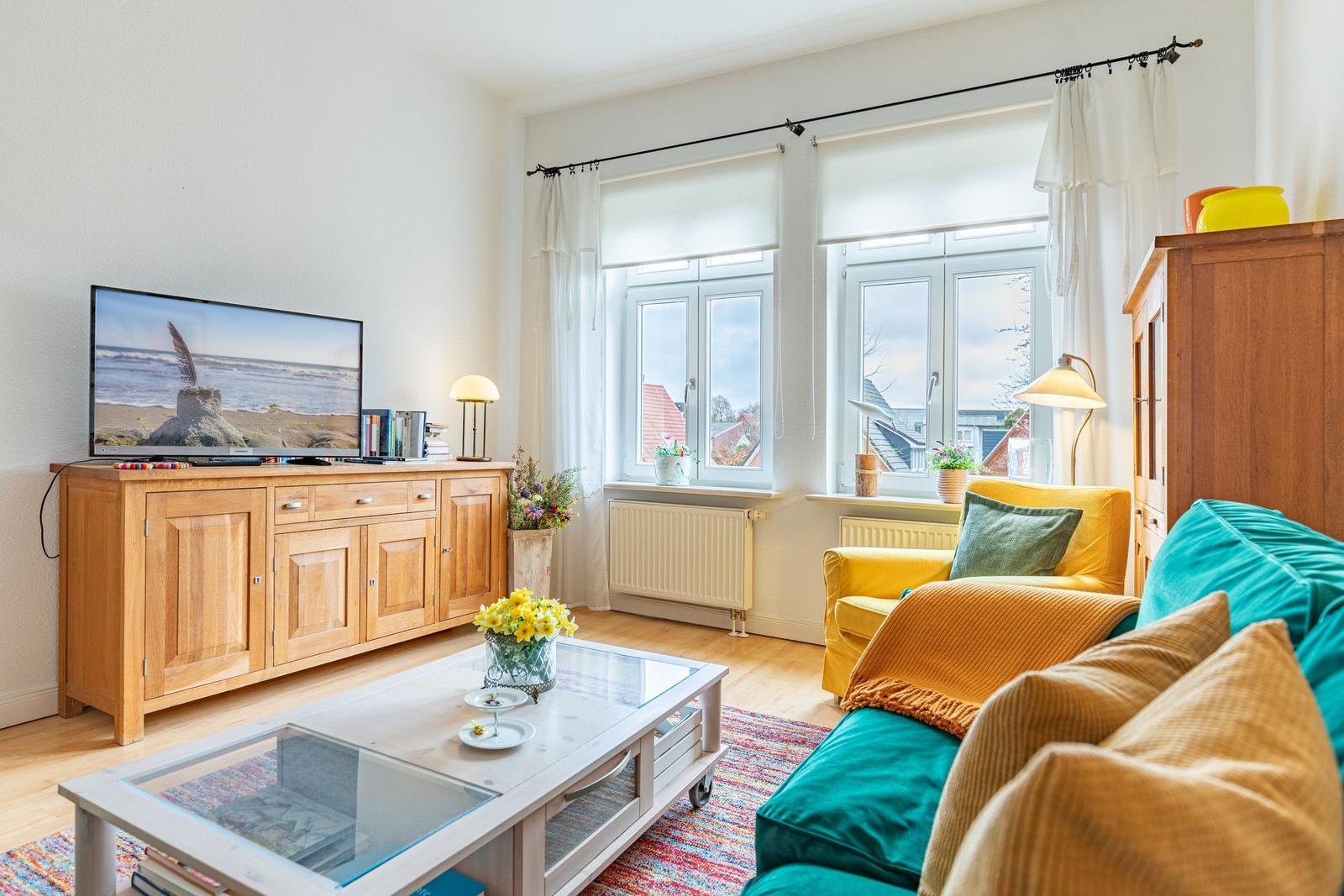 Wohnzimmer mit Fernseher, Couch und Fenstern. Holzmöbel und Blumen sichtbar.