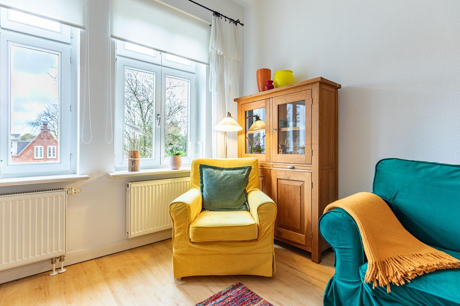 Ein Wohnzimmer mit gelbem Sessel, türkisfarbenem Sofa und Holzschrank. Fenster mit Blick auf Gebäude.