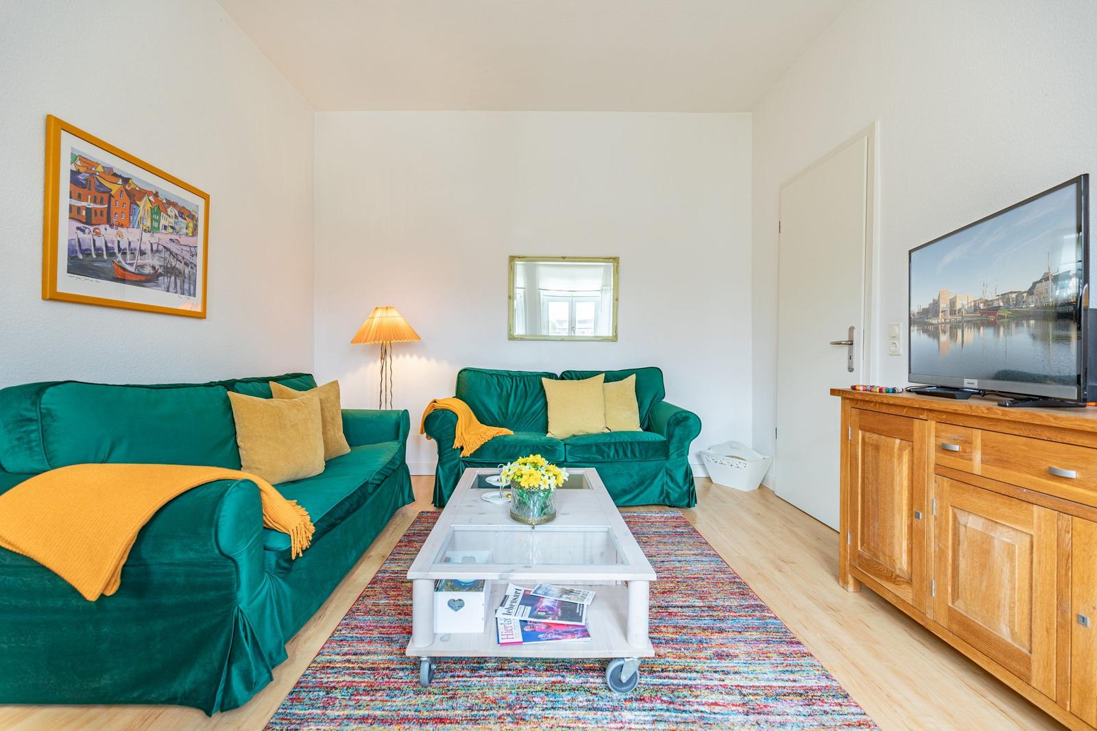 Wohnzimmer mit grünen Sofas, Holzmöbel und Fernseher. Auf dem Teppich steht ein Tisch mit Blumen.