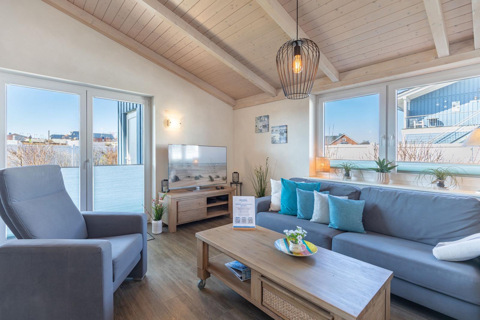 Wohnzimmer mit Sofa, Sessel und TV. Holzdecke und große Fenster.