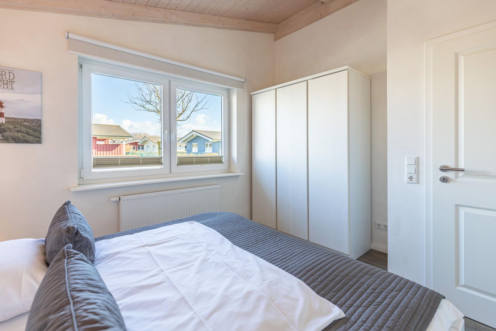 Schlafzimmer mit Bett, Kleiderschrank und Fensterblick auf Häuser.