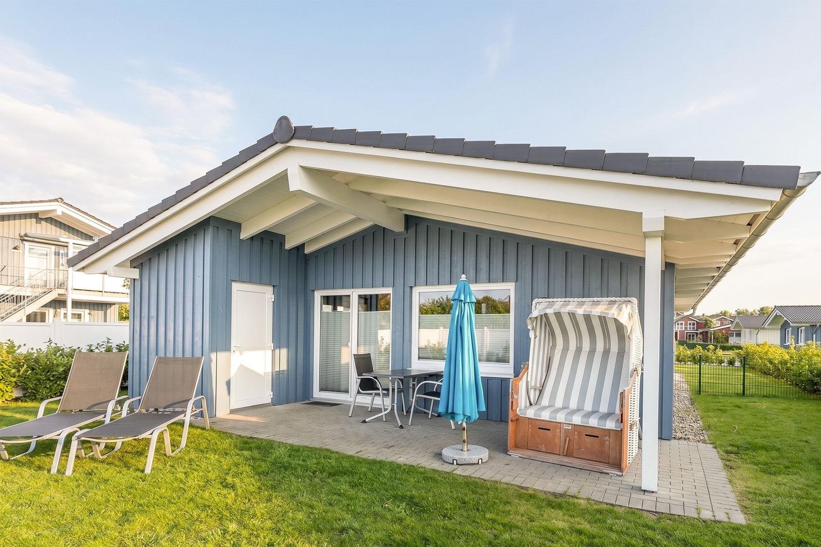 Blauer Ferienhaus mit Terrasse, Liegestühlen und Strandkorb. Grün hinter dem Haus.
