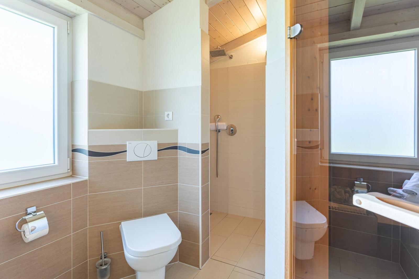 Badezimmer mit Dusche, Toilette und Waschbecken. Holzdecke und Fenster sichtbar.