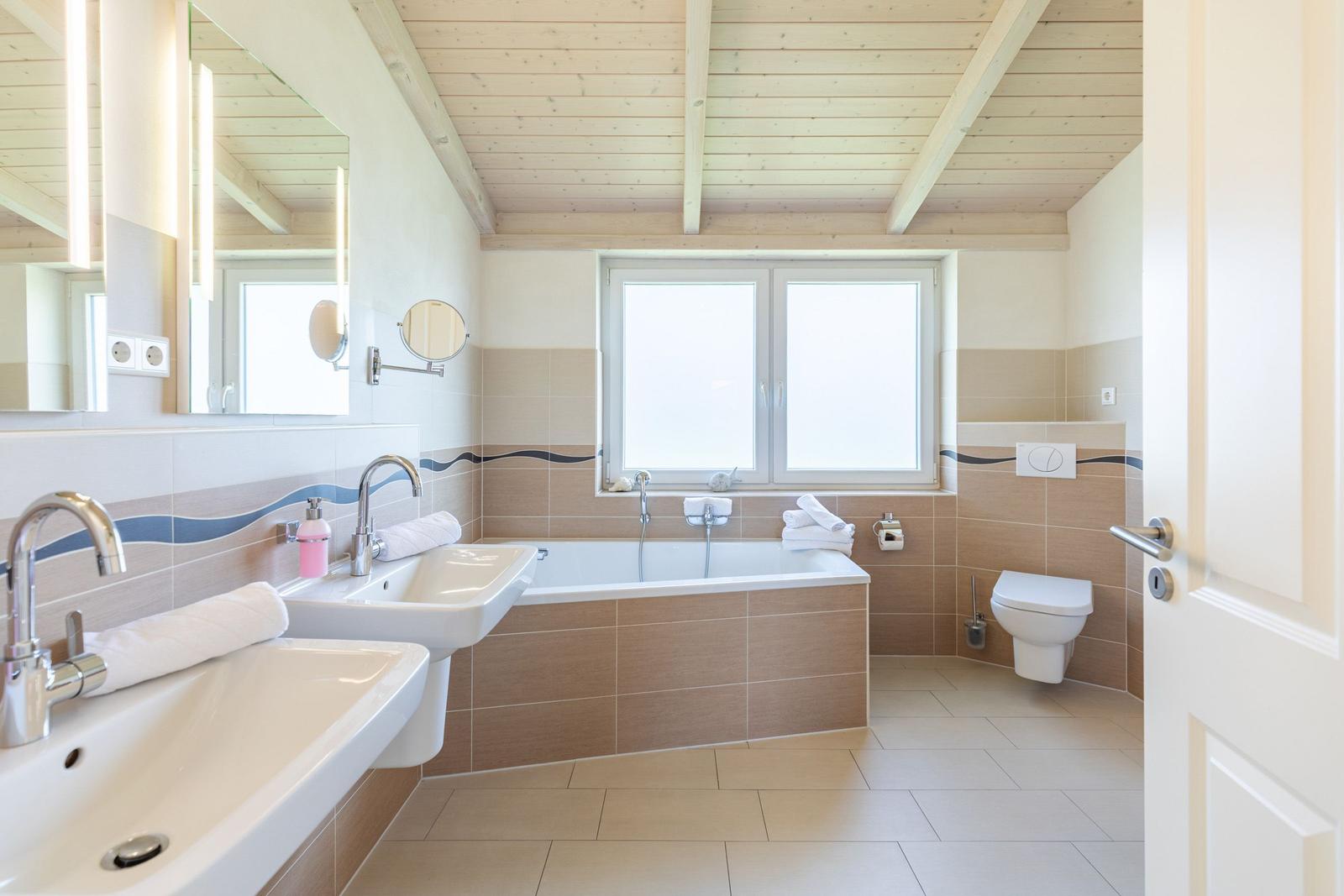 Badezimmer mit Badewanne, Doppelwaschbecken und Toilette. Holzdecke und Fenster.
