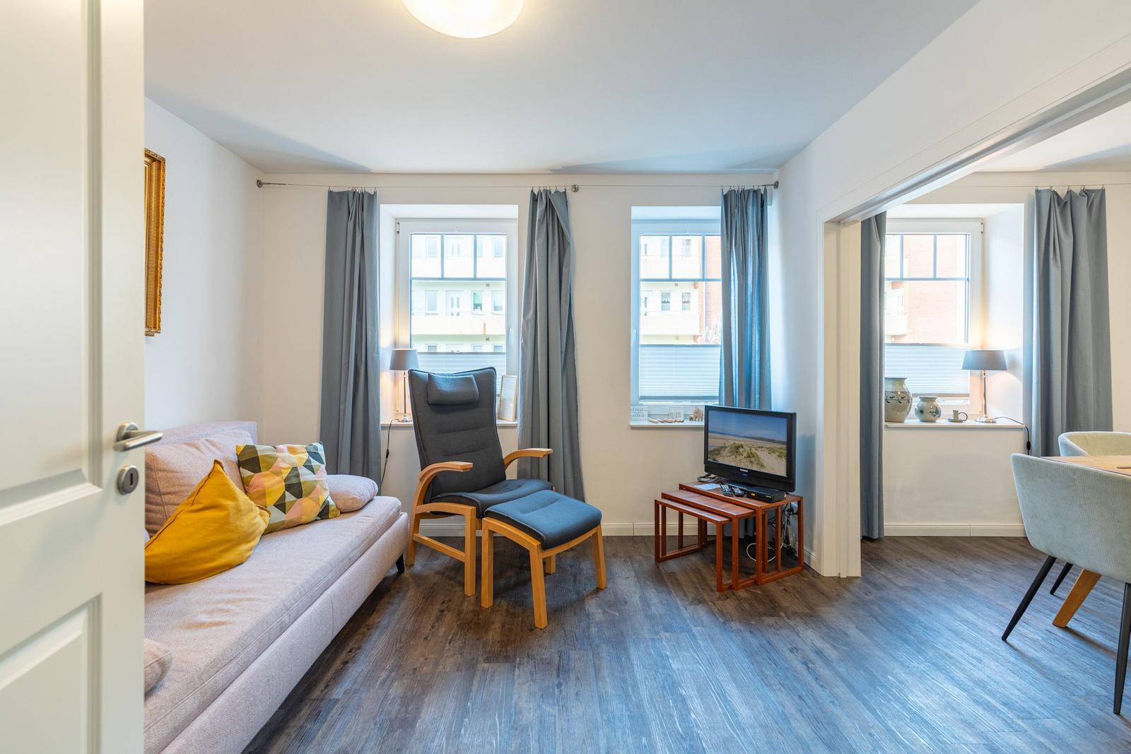 Wohnzimmer mit Sofa, Sessel, TV und Fenstern. Holzboden und graue Vorhänge.