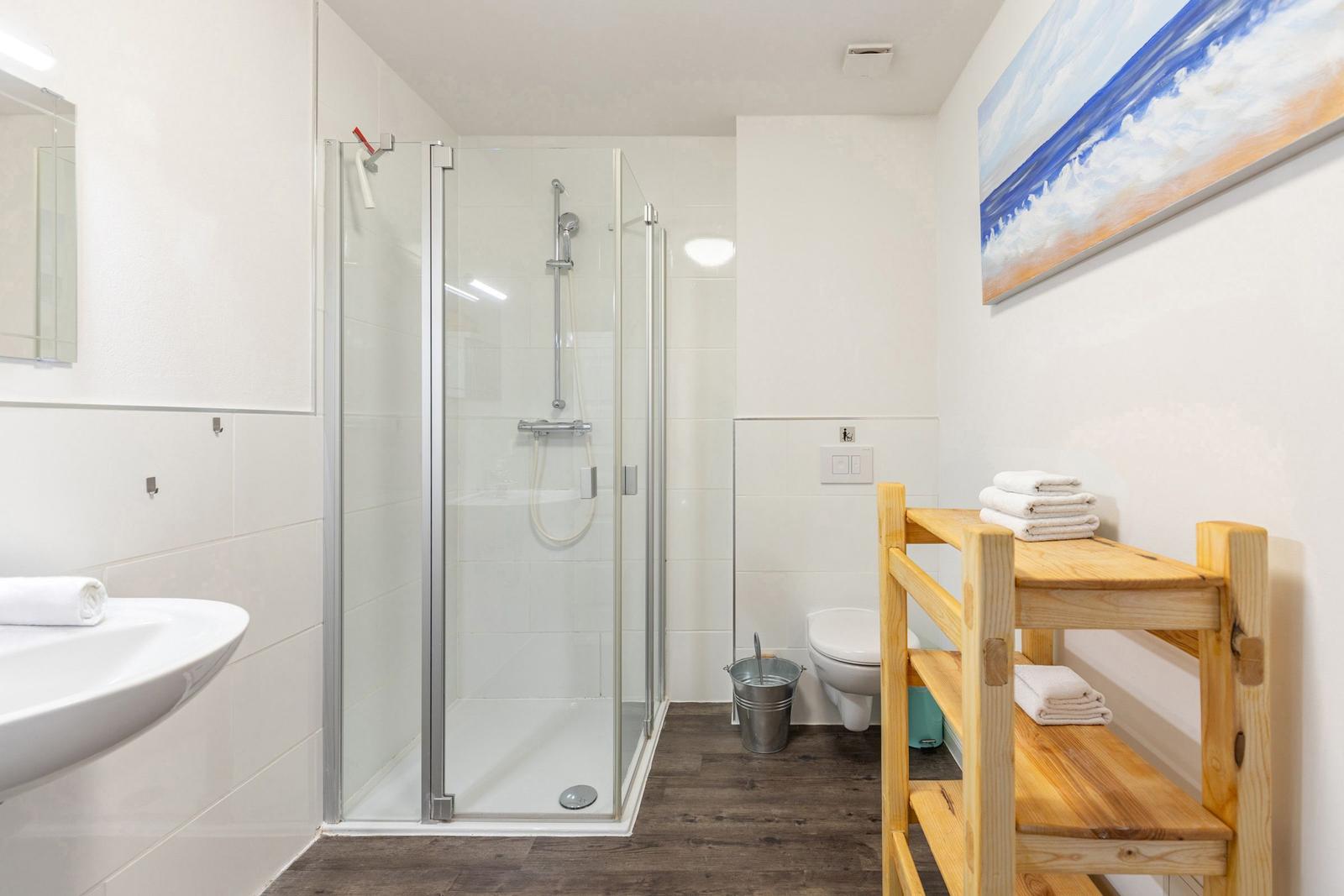 Badezimmer mit Dusche, Waschbecken und Holzregal mit Handtüchern.