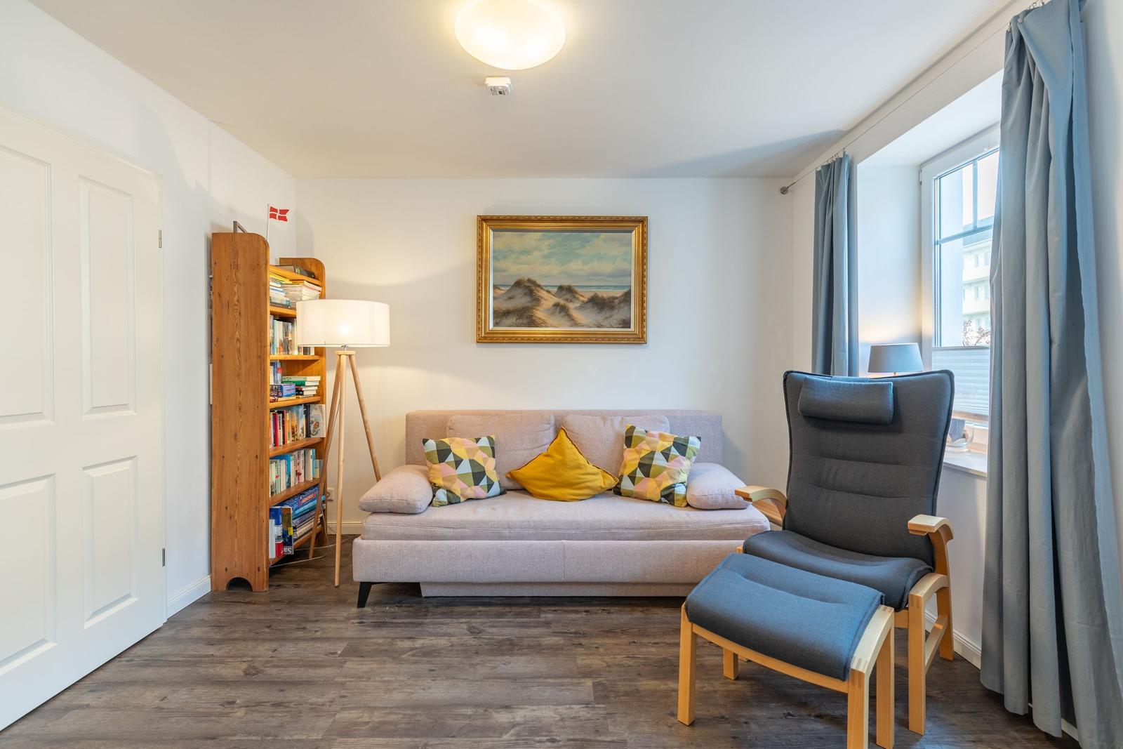 Ein Wohnzimmer mit Sofa, Sessel, Bücherregal und Fenster mit Vorhängen.