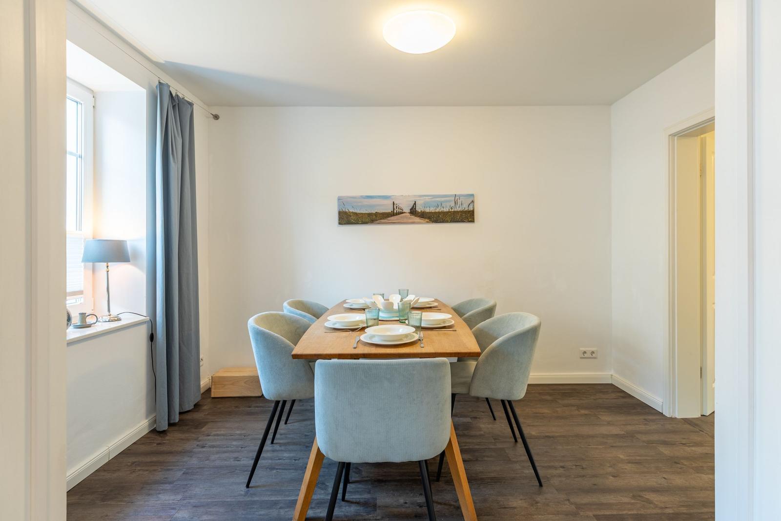 Esszimmer mit Holztisch, sechs Stühlen und Wandbild. Fenster mit Vorhang und Lampe.