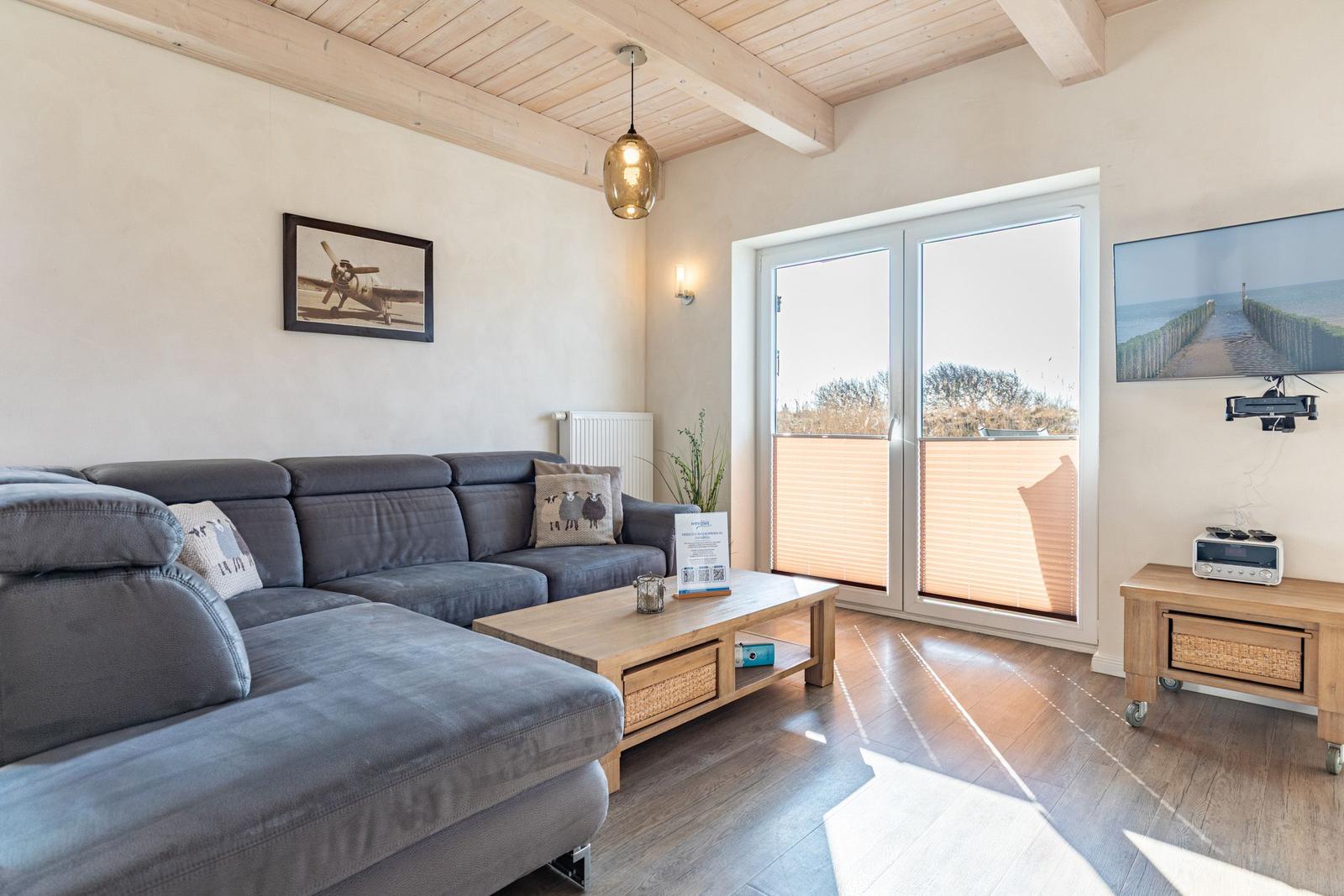 Wohnzimmer mit Sofa, Couchtisch und TV. Durchgangstür zur Terrasse mit Blick auf den Strand.