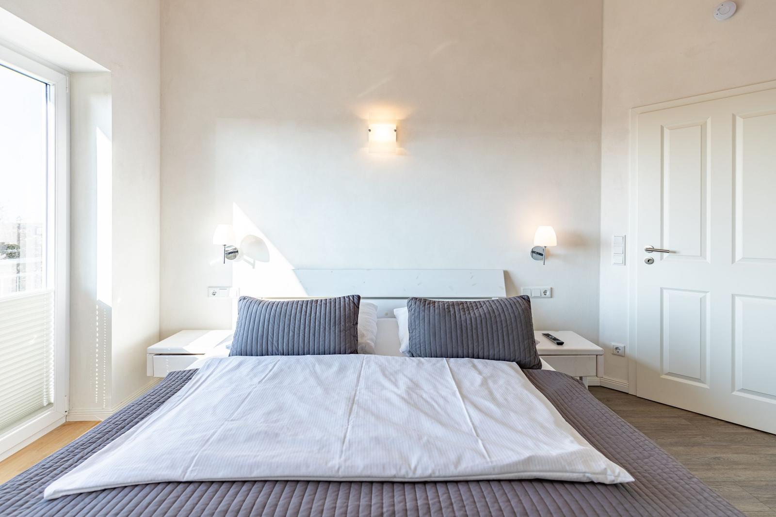 Schlafzimmer mit Doppelbett, zwei Nachttischen und Wandlampen. Fenster mit Jalousie und Tür.