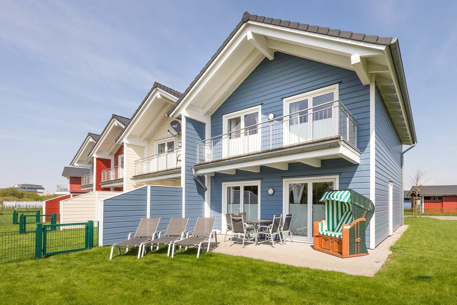 Blau gestrichenes Ferienhaus mit Balkon und Gartenbereich mit Liegen und Strandkorb.