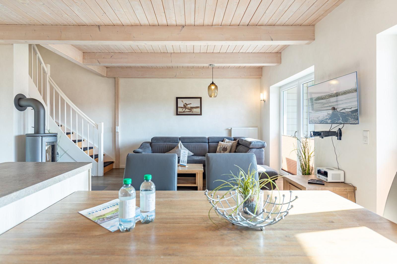 Wohn- und Essbereich mit Sofa, TV und Holzdecke. Tisch mit Wasserflaschen und Pflanze.
