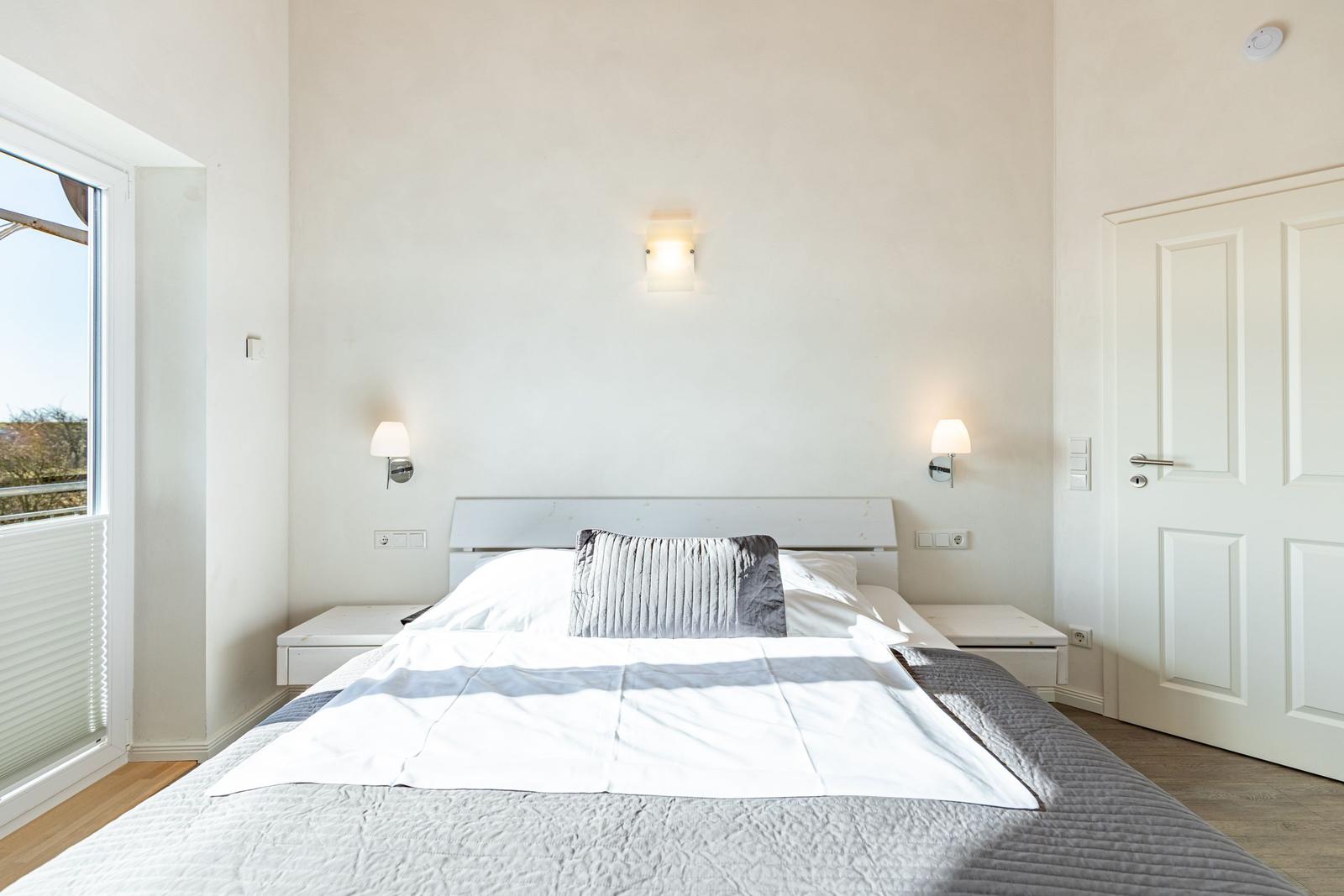 Schlafzimmer mit Bett, zwei Nachttischen und Wandlampen. Fenster mit Jalousie und Tür.