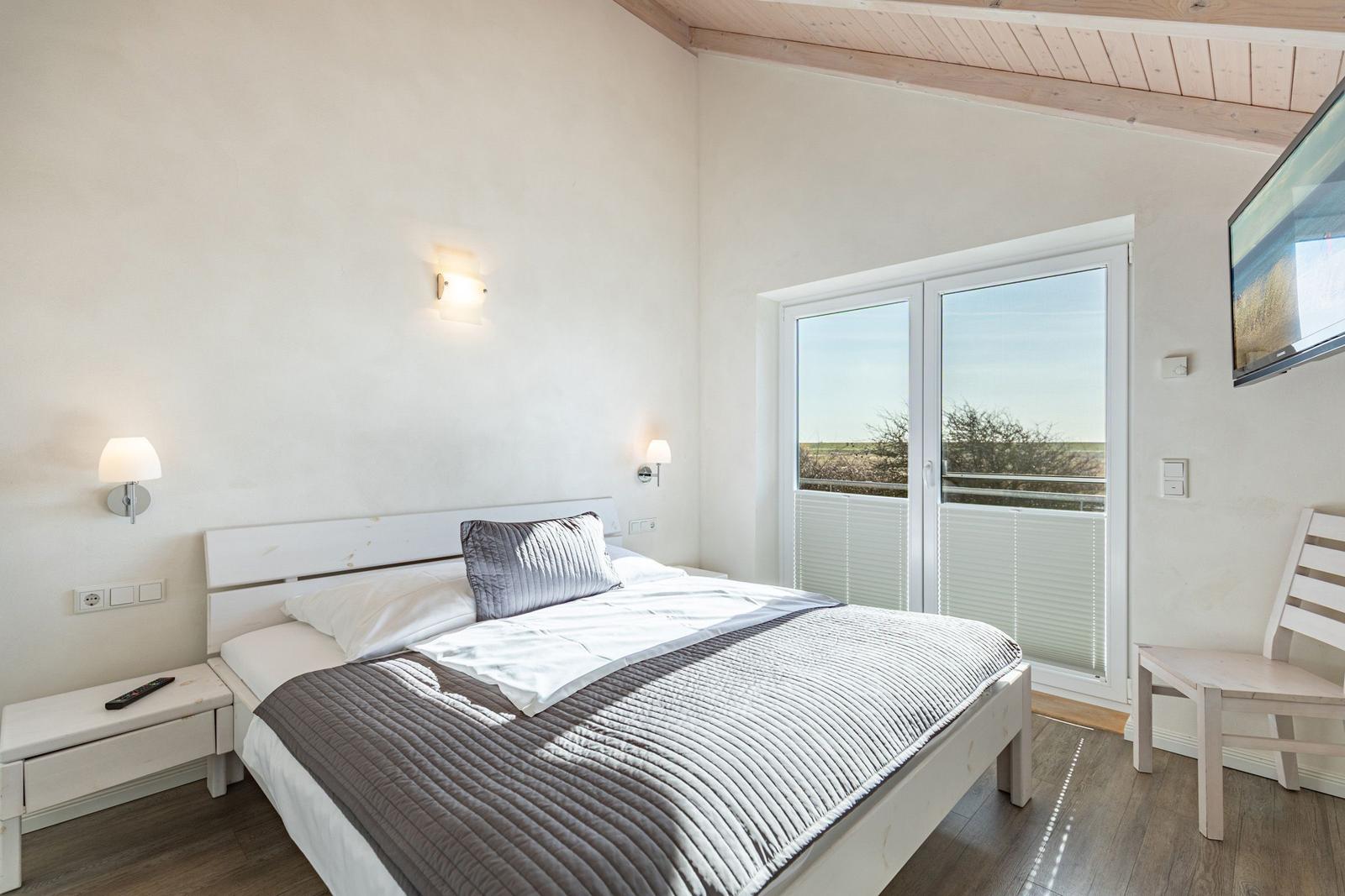 Schlafzimmer mit Bett, Schrank und Balkontür. Wandlampen und Fernseher an der Wand.