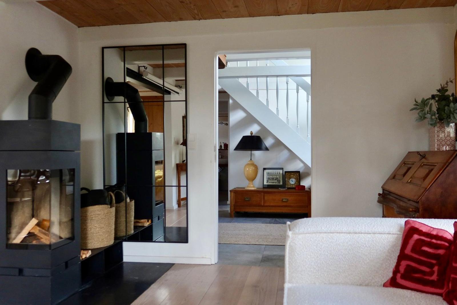 Wohnzimmer mit Kamin, Spiegel und Sitzgelegenheit. Treppenhaus im Hintergrund.