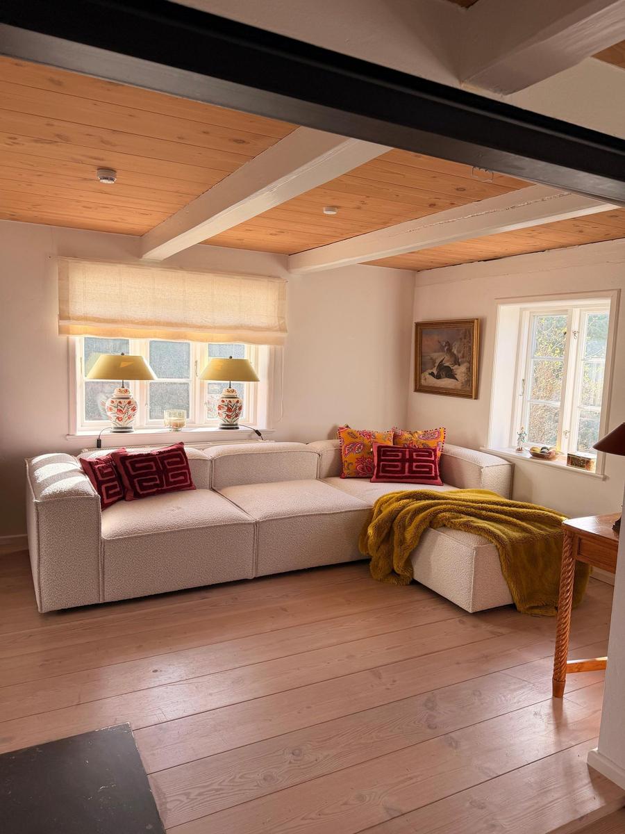Wohnzimmer mit L-förmigem Sofa, Holzboden und Deckenbalken. Fenster mit Lampen und Kissen.