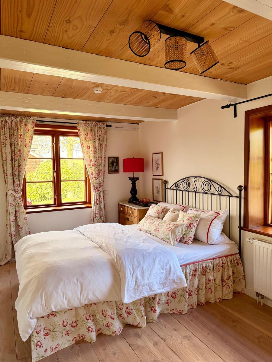 Schlafzimmer mit Bett, Holzboden und Fenster mit floralen Vorhängen.