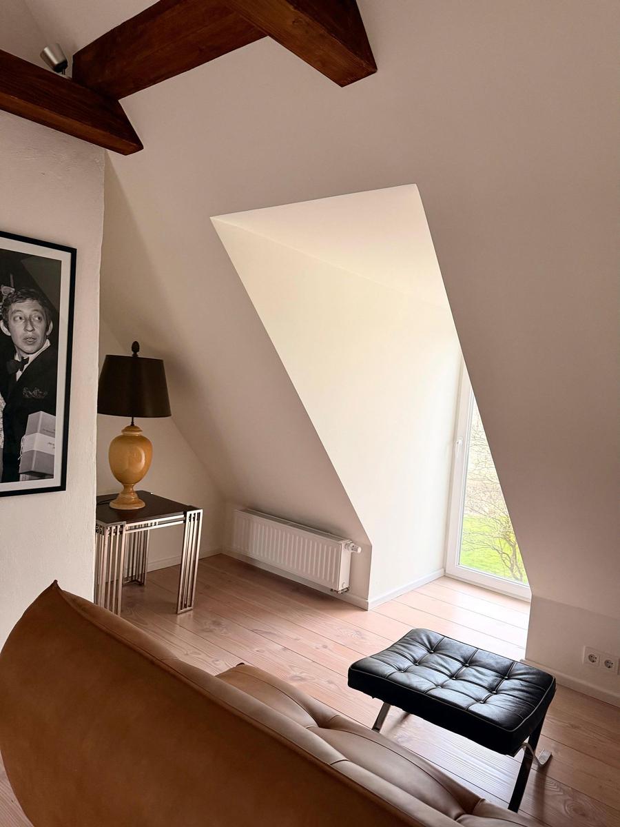 Dachgeschosszimmer mit Holzboden, schwarzem Sitzhocker und schrägem Fenster.