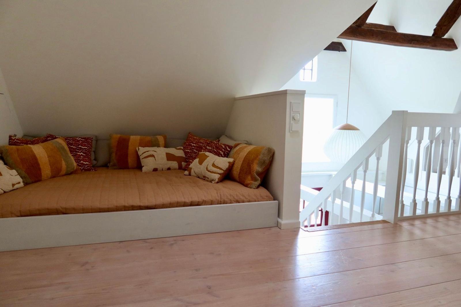 Ein Bett mit Kissen unter einem Dachboden, neben einer Treppe mit weißem Geländer.