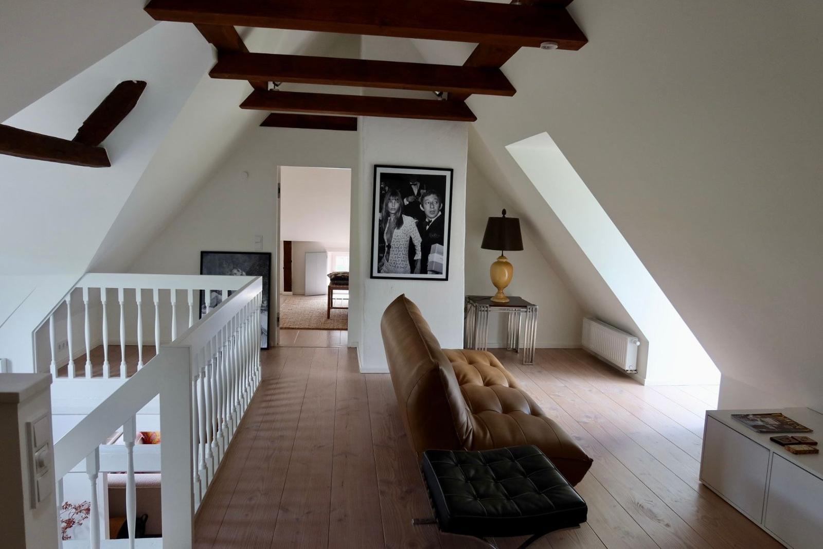Dachgeschosswohnung mit Holzbalken, Sofa und Treppe. Lichtdurchfluteter Raum mit Fenster.